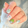 Hand with intricately designed nail green and white St. Patrick's Day nail wraps featuring lucky charms and plaid, held against a neutral background.