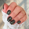 Hand with black and green lucky clover nail wrap design wearing a white textured sleeve.