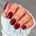 Hand wearing Garnet Glitz burgundy nail wraps with fine dark red glitter and a glossy finish, beside a white knit sleeve.