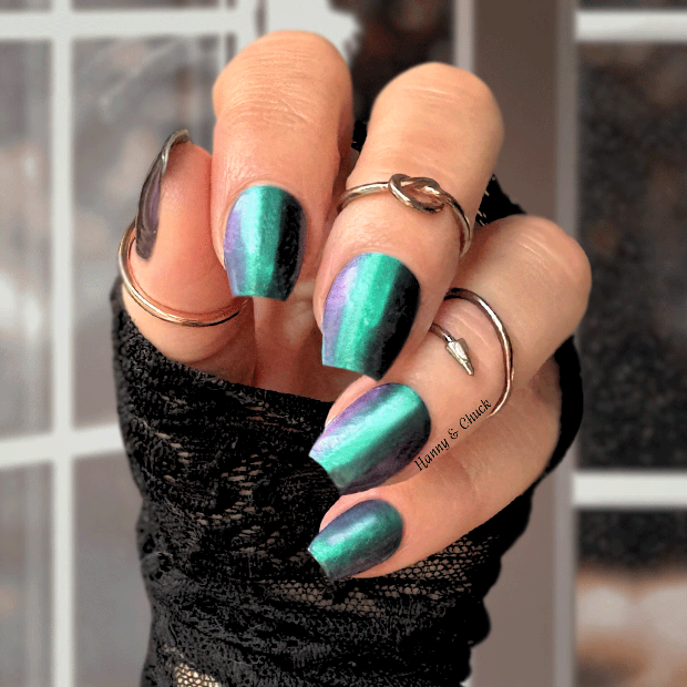 Hand with green and purple colour shift nail polish wraps wearing multiple rings against a blurred window background