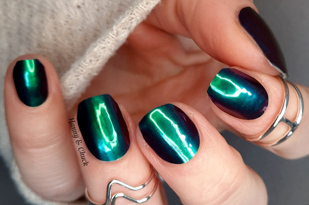 A person's hand with fingernails wrapped in a green to purple color-shifting nail wrap, wearing a silver ring, and partially covered by a beige knitted sweater.
