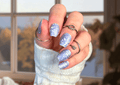 Hand with glittery blue nail polish wraps wearing a white sleeve and rings.