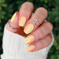 A hand with Pineapple Sunrise nail wraps showing vibrant yellow nail polish. The hand is also adorned with silver rings, and the background consists of green foliage.