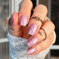 Hand with glittery pink nail wraps wearing multiple rings against a blurred background