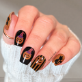 Halloween-themed nail wrap, featuring black nails decorated with glittery orange pumpkins, black cats, and purple witch hats, worn with a white knit sweater.