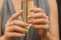 Close-up of hands holding a champagne flute with glittery gold nail wraps against a neutral background