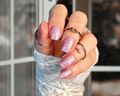 Hand with glittery pink nail wraps wearing multiple rings against a blurred background