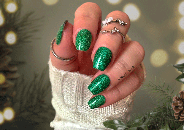 Hand with green glittery nail stickers wearing a white textured sleeve against a blurred natural background.