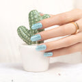 Hand with glittery blue nail polish wraps holding a small potted cactus.