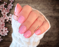 Hand with pink nail polish wraps holding a pink lipstick on a wooden background with cherry blossoms.