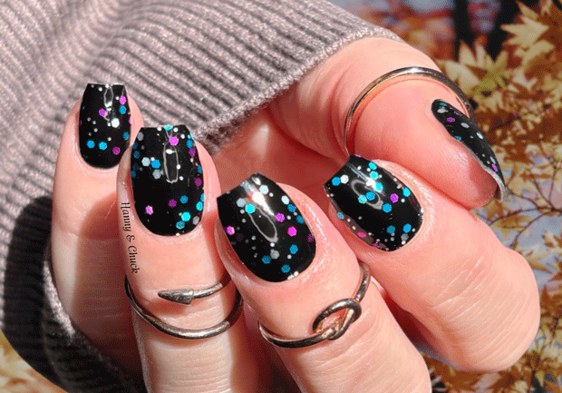Hand with black nail wraps with colorful purple, blue and silver glitter dots against a outdoor background.