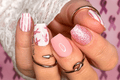 Close-up of a hand with pink and white nail polish wraps, wearing multiple rings on a pink background.