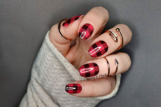 A person's hand with fingernails adorned with red and black plaid pattern nail wraps, wearing a grey sweater and gold rings on the fingers.