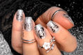 Silver nail wraps with intricate designs on a hand wearing multiple rings.