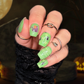 Hand with green Halloween-themed nail wraps against a dark background with pumpkins.