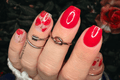 Close-up of a hand with red nail polish wraps and floral poppy designs, wearing rings, against a blurred floral background.