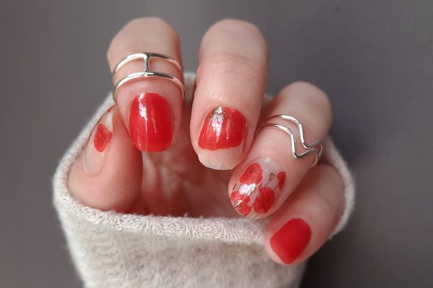A hand with red nail polish and a sheer red nail wrap with poppy flower accents.