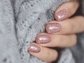 A close-up of a person's fingernails with light pink glitter nail wraps, holding a textured grey fabric.