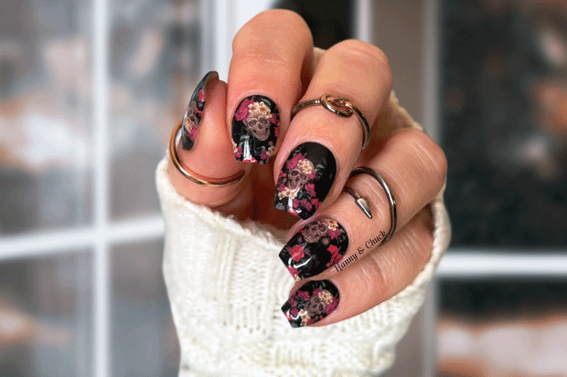 Hand with floral skull nail wrap design wearing a white sleeve and rings.