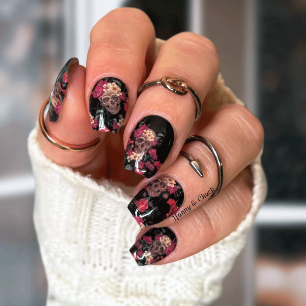 Hand with floral skulls nail wrap design wearing a white sleeve and rings.