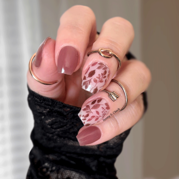 Hand with pink nail stickers and floral design, wearing multiple rings, against a neutral background.