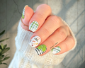 Hand with intricately designed nail green and white St. Patrick's Day nail wraps featuring lucky charms and plaid, held against a neutral background.