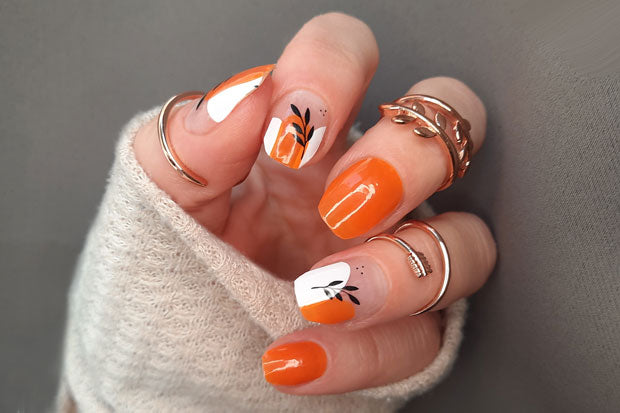 A hand with orange and white nail wraps adorned with black leaf accents, wearing silver rings on each finger.