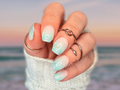 Hand with turquoise and coral gradient nail wraps wearing rings, blurred ocean background