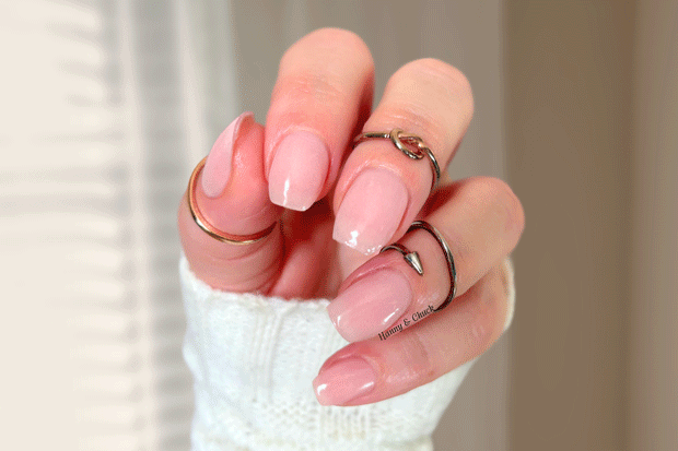 Hand with sheer light pink nail polish wraps and multiple rings on a neutral background