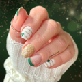 A hand showcasing nail polish wraps with green French tips, white and gold horizontal stripes, and glitter accents, with a shamrock design.