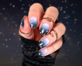 A close-up image of a hand with fingernails painted with a celestial design nail wraps, featuring a gradient from lavender pink to black, with white sparkles and holographic glitter, and crescent moon accents.