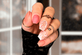 Hand with pink and coral nail polish wraps with floral designs, wearing rings, against a blurred background.