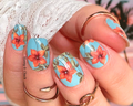 A photo of a person's hand showcasing nail wraps with a vibrant orange lilies pattern on a deep teal background.