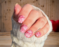 Hand with pink valentines day nail design wearing a white sleeve against a wooden background