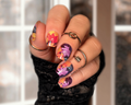 Hand with colorful maple leaf nail art stickers wearing black fingerless gloves against a blurred indoor background