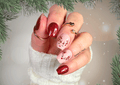 Hand with festive nail art featuring red and pink reindeer design nail wraps, surrounded by Christmas decorations.