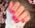 Hand with sheer pink nail polish wraps and hearts and bows designs, wearing a ring, against a blurred floral background.