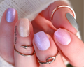A close-up image of a person's fingernails, which are adorned with a gradient nail polish wrap that transitions through colors of pink, purple, beige, and blue. The nails are also fitted with gold-colored nail rings.