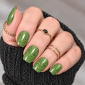 A Little Envy apple-green glitter nail wraps with golden sparkle, shown on a hand wearing gold rings and a black knit sweater.