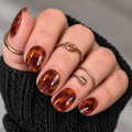 Glossy amber and dark brown tortoiseshell nail wraps on short rounded nails, shown with gold rings and a black knit sleeve.