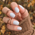 Freshly Fallen white nail wraps with fine silver glitter on short nails, styled with gold rings and a brown knitted sleeve against blurred autumn foliage.