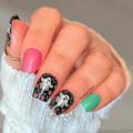 Close-up of manicured nails featuring the Ghostly Garden design: pink, mint green, and black nail wraps with white ghost and pink floral details, displayed on a white knit sweater