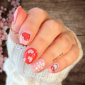Hand with pink and red nail stickers on a wooden surface with cherry blossoms.