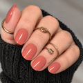 Hand wearing Honey Persimmon coral terracotta nail wraps with a glossy finish, gold rings and a black knit sleeve.