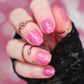 A hand with hot pink nail polish and heart patterns, wearing gold rings, against a pink glittery background.