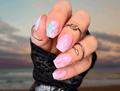 Hand with pink and blue nail polish stickers against a blurred beach sunset background
