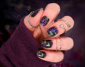 Hand with purple and green glittery nail polish wearing silver rings on a dark background