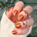 A hand with burnt orange glitter nail wraps and multiple rings, positioned on a green wooden surface with white flowers in the background.