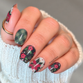Close-up of hand wearing Christmas nail wraps with green plaid, black floral, and white poinsettia nail designs.