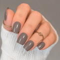 Close-up of glossy warm taupe-grey nail wraps on short square nails, styled with gold rings and a white knit sleeve.